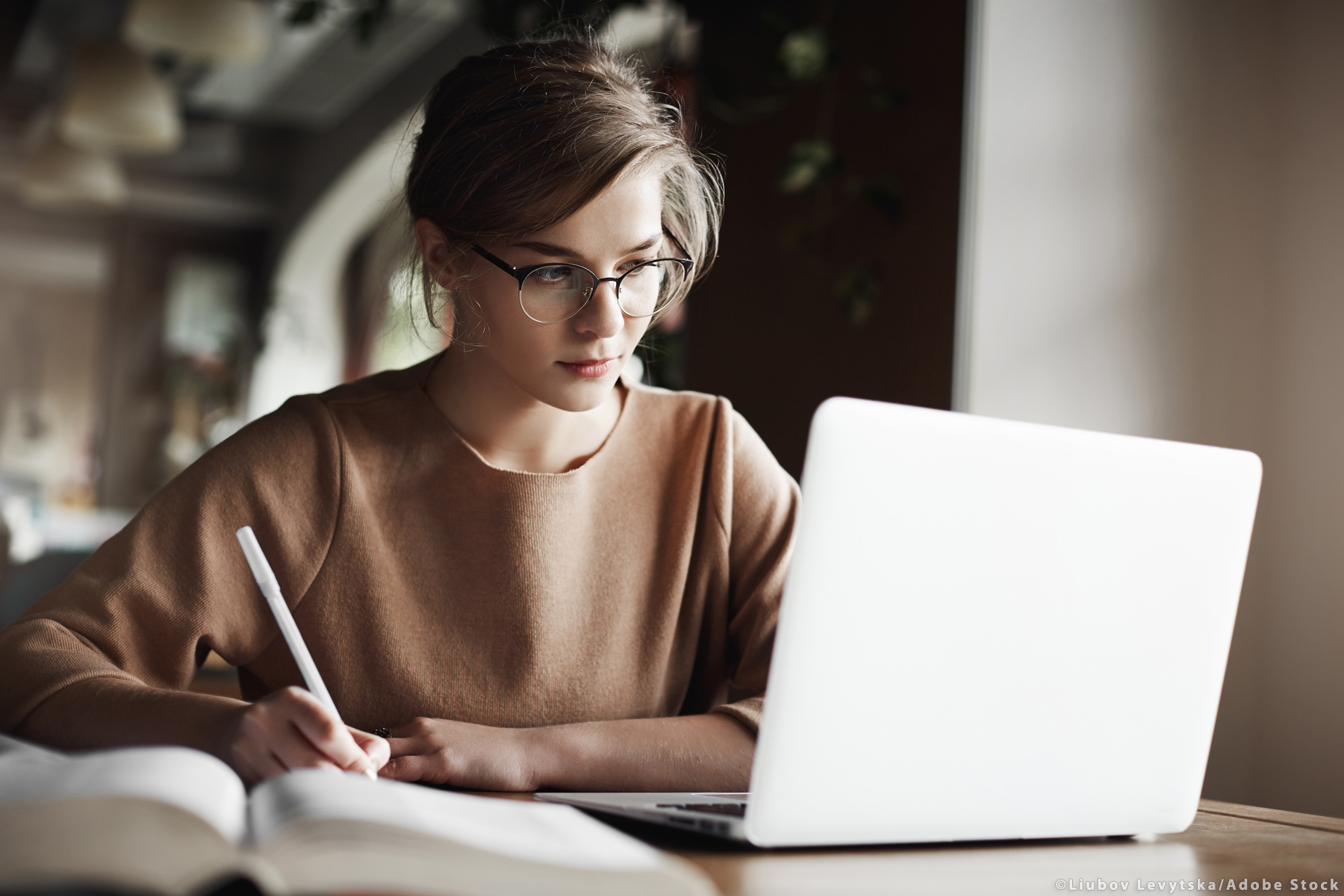 Creative good-looking european female with fair hair in trendy glasses, making notes while looking at laptop screen, working or preparing for business meeting, being focused and hardworking,