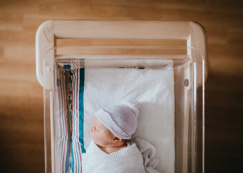 A baby just born at the hospital rests in a hospital bassinet crib, wrapped in a swaddle and wearing a beanie hat.