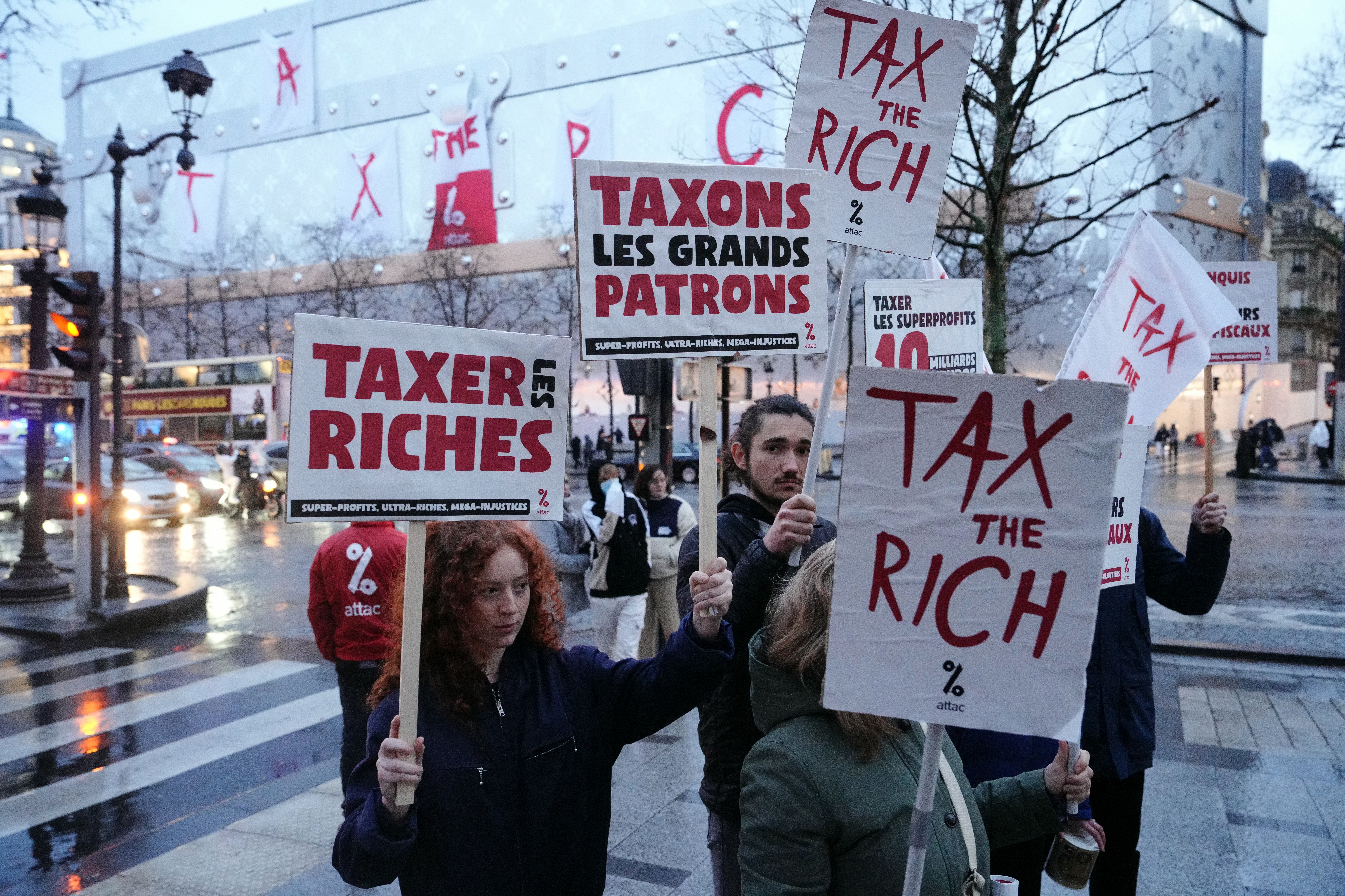 Protesters hold placards reading "Tax the rich" as banners are displayed on the rooftop of the new Louis Vuitton hotel under construction concealed in a Louis Vuitton trunk during an action by Attac France NGO on the Champs Elysees in Paris on February 24, 2024. Several dozen Attac activists successfully unfurled a giant "Tax the Rich" banner on February 24, 2024 from the top of the facade of luxury group LVMH's future hotel on the Champs-Elysees in Paris, AFP journalists observed. (Photo by Dimitar DILKOFF / AFP)